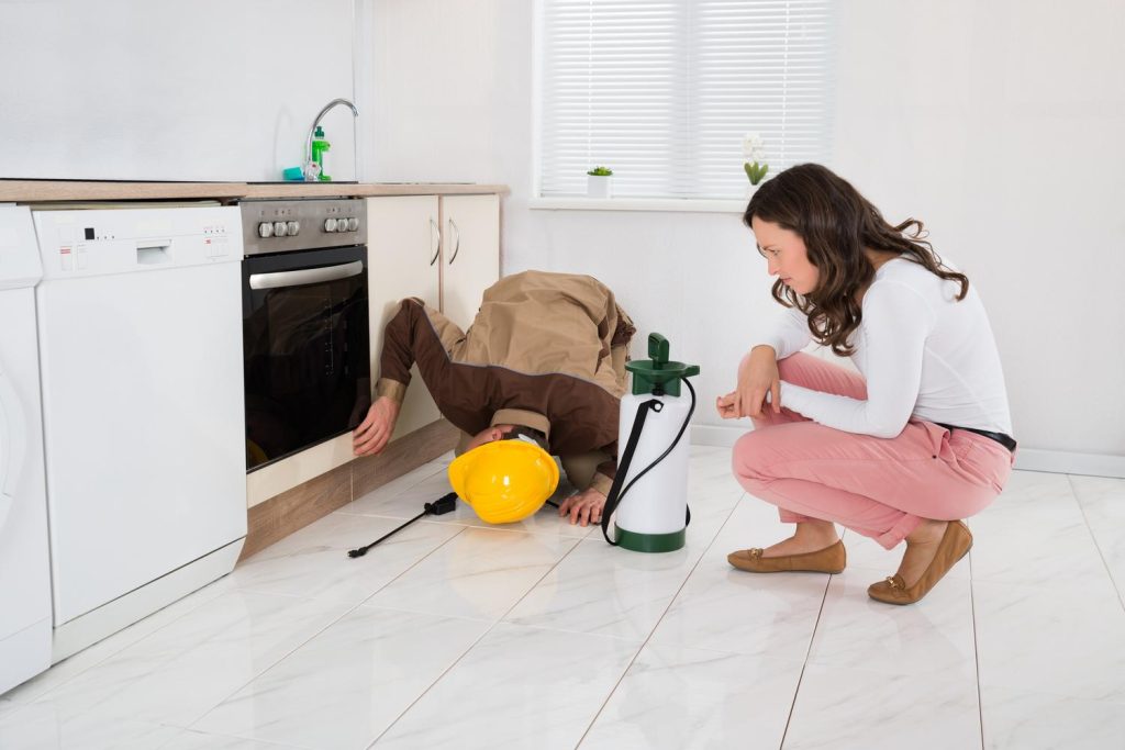 Man wearing a yellow safety hat doing pest control in the kitchen while a girl watches